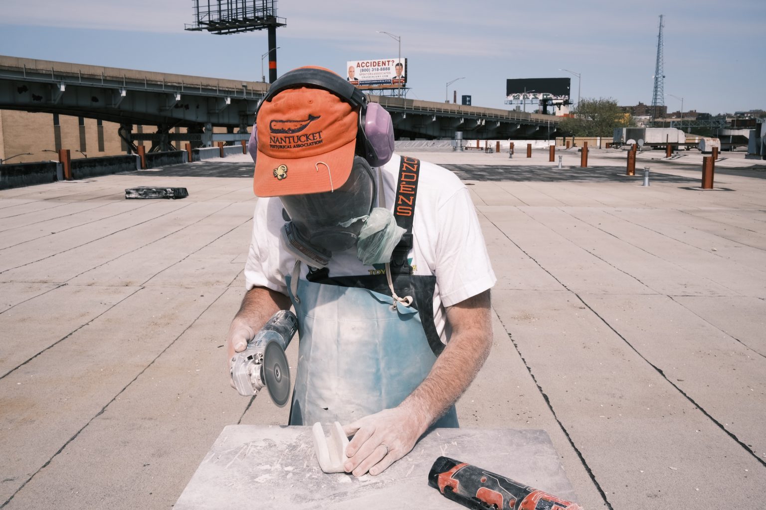 Colour photograph of a person stone sculpting on a rooftop, wearing an orange Nantucket cap, purple earmuffs, white shirt and protective gear while working with a handheld angle grinder on concrete.